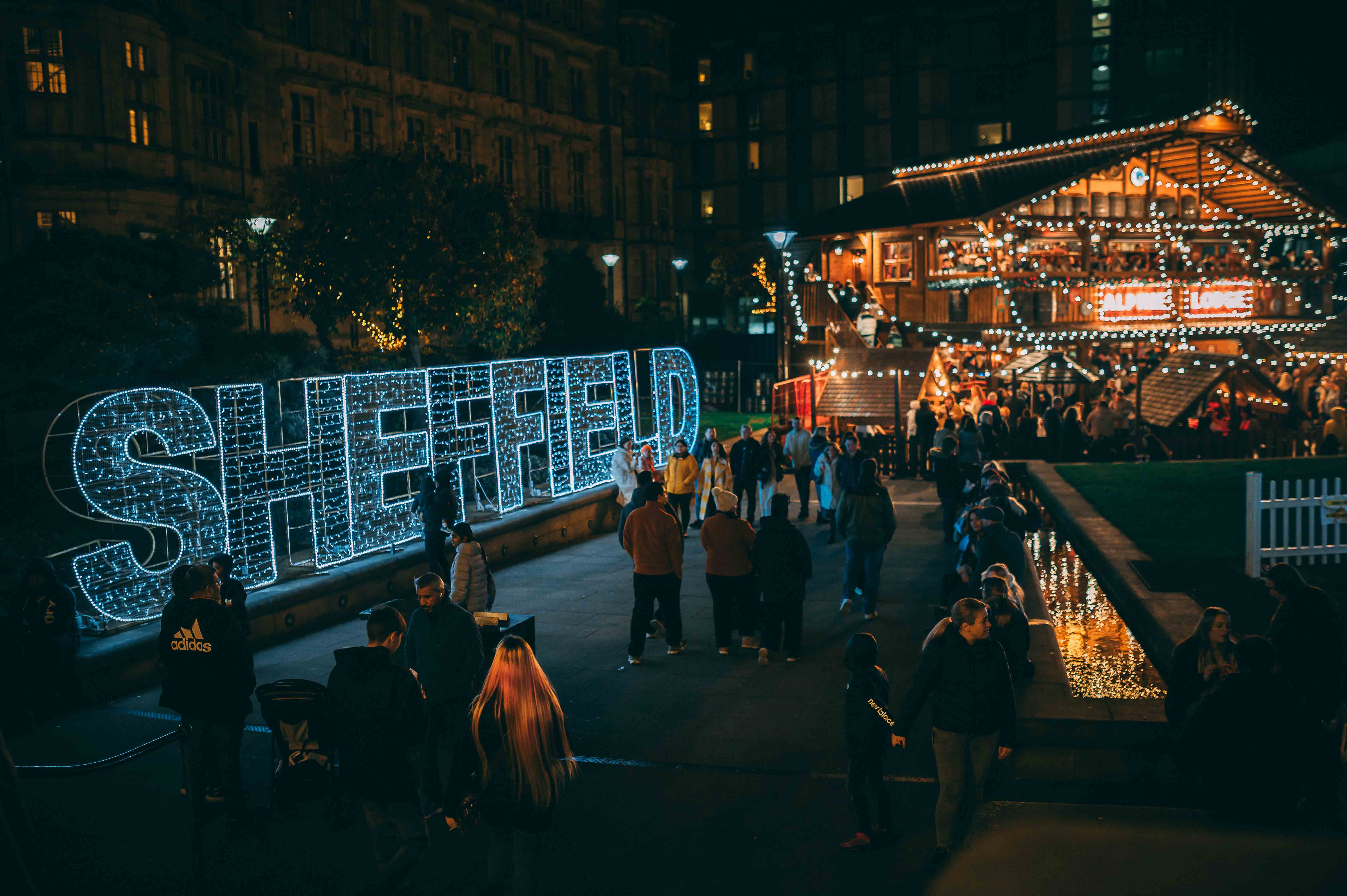 Illuminated Sheffield sign at night positioned alongside the wall of the Peace Gradens in Sheffield with the twinkly lights of a large Alpine Lodge visible to the side in the background and people wandering through 