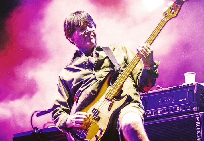 A musician is performing on stage, playing an electric bass guitar. The person is wearing a long-sleeved shirt and shorts, with the guitar strap across the shoulder. The background is filled with vibrant pink and purple stage lighting, and there is audio equipment and a cup placed on top of an amplifier next to the performer.
