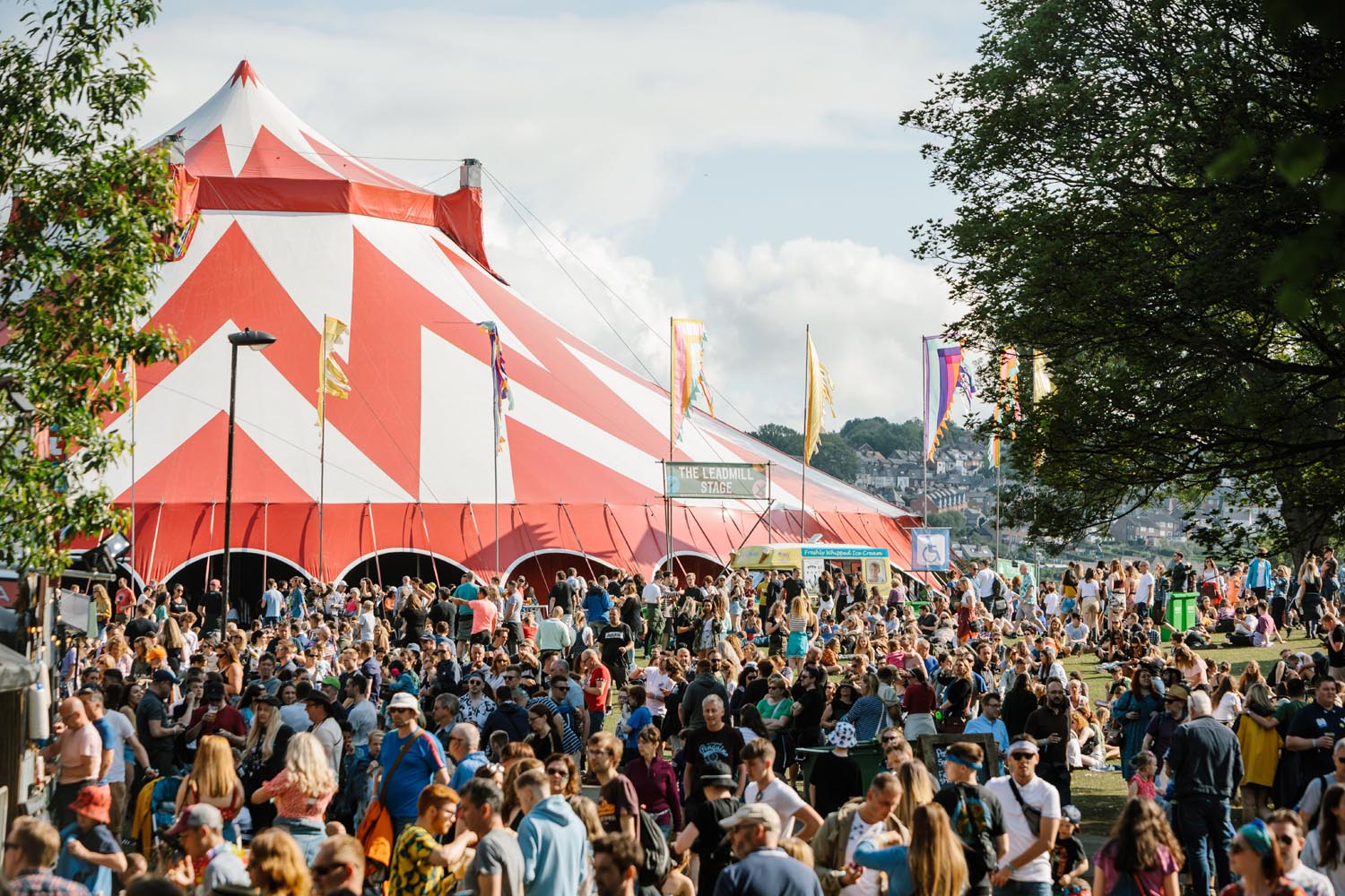 Large outdoor festival crowd gathered near a big red-and-white striped tent, with colourful flags and trees in the background under a partly cloudy sky.