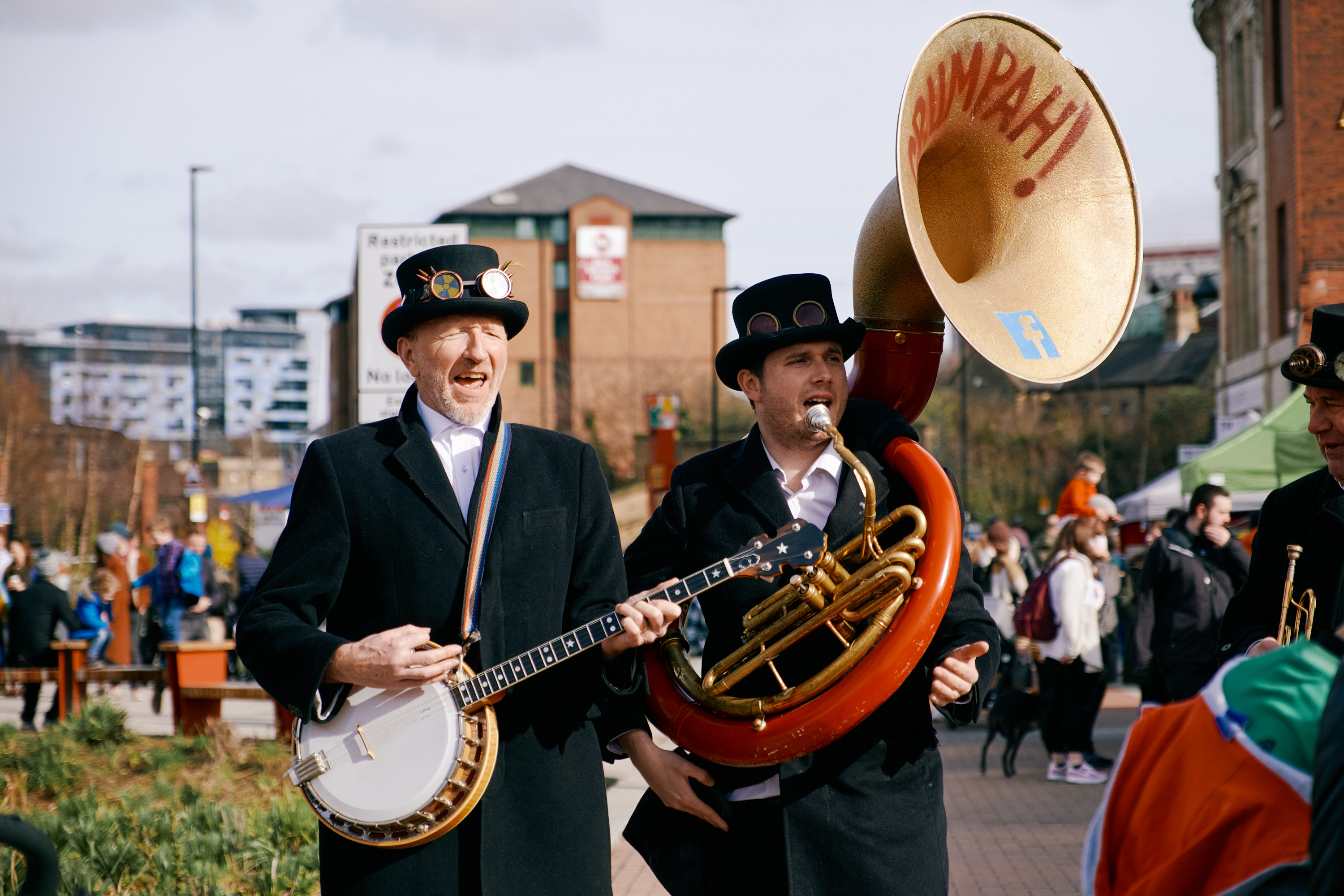 Two musicians, one playing a banjo and the other a brass sousaphone, perform at an outdoor market in Sheffield.
