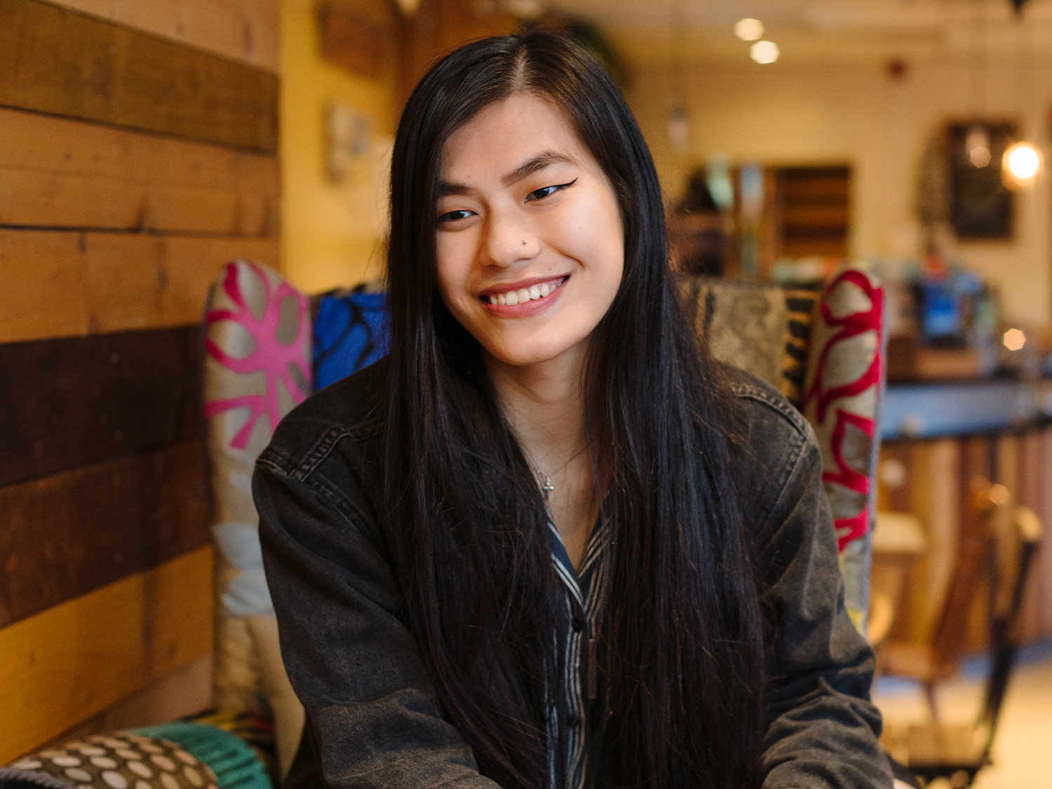 A person with long straight dark hair is seated indoors on a patterned chair. The individual is wearing a dark denim jacket over a striped shirt. The background features wooden paneling on the wall and warm ambient lighting, suggesting a cozy café or restaurant setting.