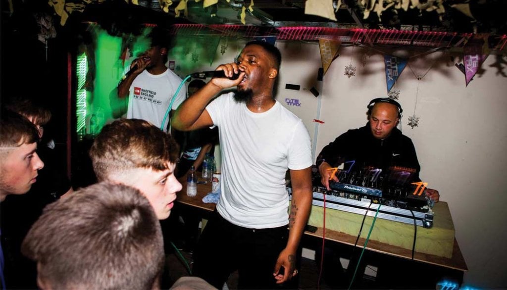 A man sings into a microphone in front of a crowd. Behind him is a man mixing on two DJ decks.