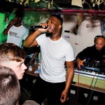 A man sings into a microphone in front of a crowd. Behind him is a man mixing on two DJ decks.