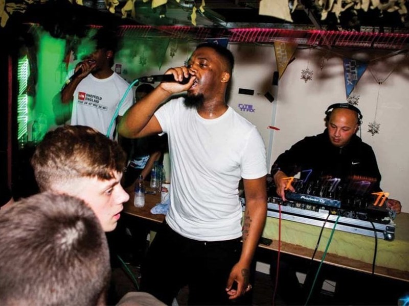 A man sings into a microphone in front of a crowd. Behind him is a man mixing on two DJ decks.