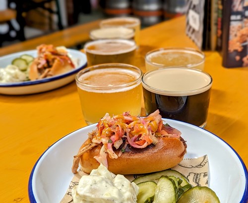 Close-up of a plate with a loaded hot dog topped with shredded meat and pickled vegetables, served with sliced pickles and a dollop of creamy sauce. In the background, there are additional plates and a flight of four craft beers on a bright yellow table.