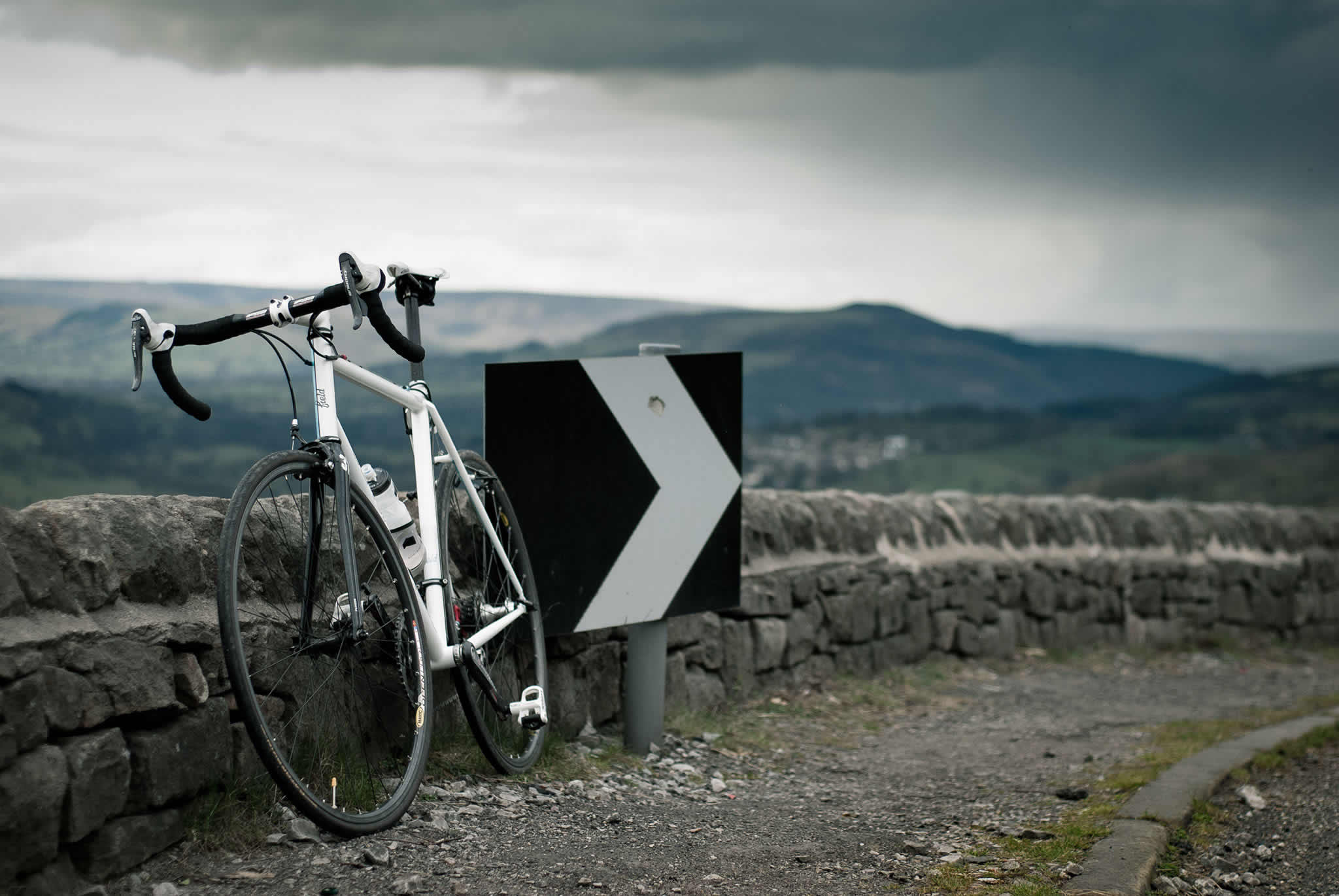 A racing bike leaning against a stone wall at the edge of a road out in the countryside.