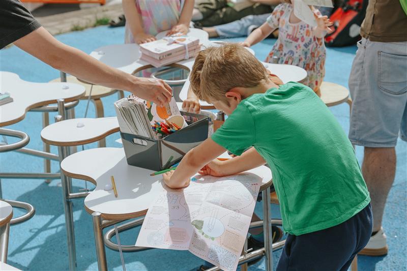 Child leaning over a table with a coloring sheet, surrounded by other children and adults at an outdoor craft activity. The table holds a box of colored pencils and markers, and several stools are arranged on a bright blue surface.