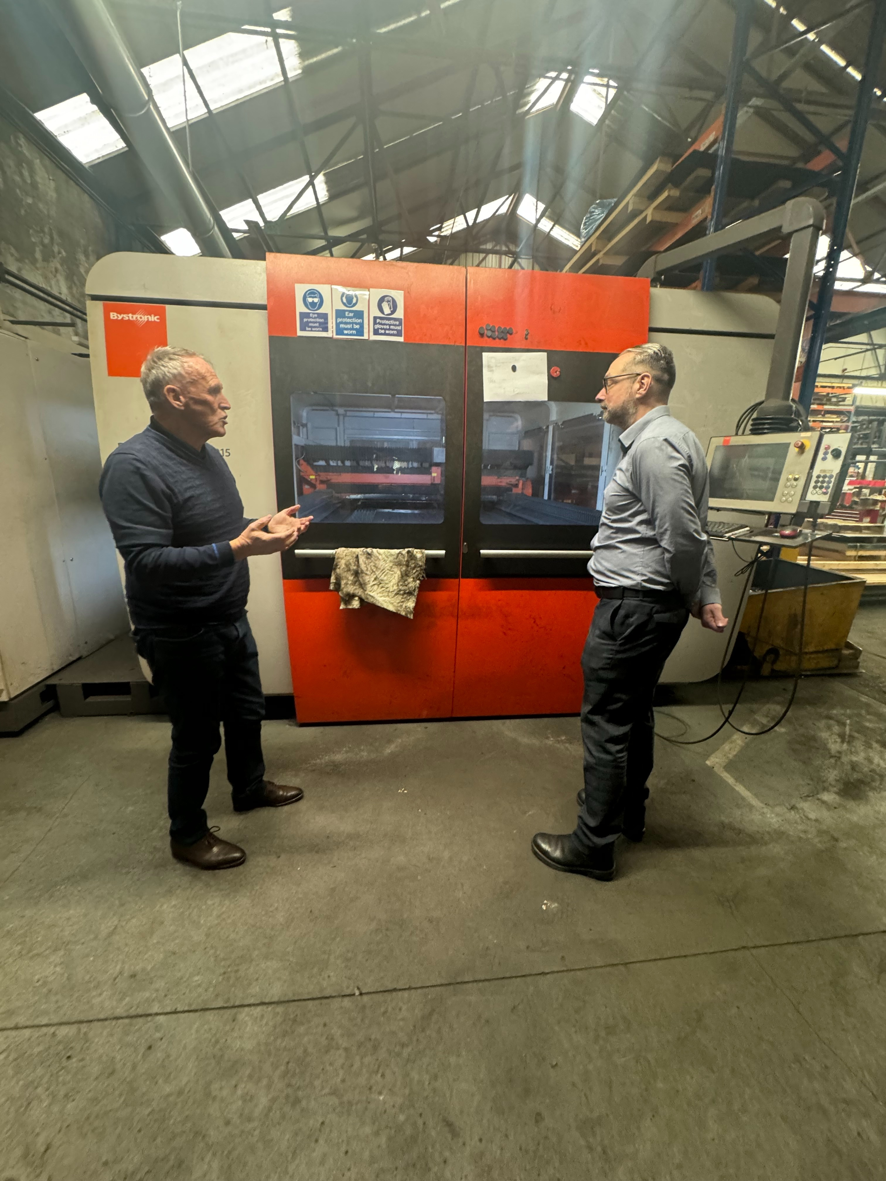 Two people standing in front of a large industrial machine with a bright orange and black exterior in a factory setting. One person is gesturing with hands while the other stands near a control panel attached to the machine. The background shows metal beams, shelves, and equipment under a high roof with skylights.