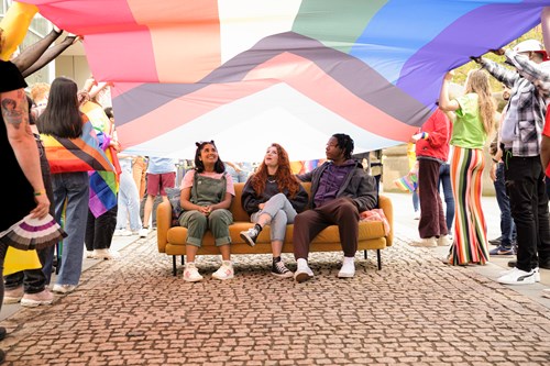 Three people sitting on a yellow sofa under a large Progress Pride flag being held overhead by a group of people. The flag displays rainbow colors along with chevron stripes in black, brown, white, pink, blue, and light blue. The scene takes place outdoors on a cobblestone walkway, with individuals standing around holding sections of the flag.