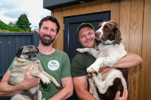 Two men, standing outside a wooden building, are holding a dog each and smiling.