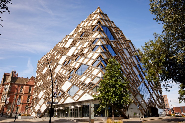 Modern pyramid-shaped building with a striking geometric design featuring glass panels and wooden lattice framework, located on a street corner with trees and older brick buildings nearby under a clear blue sky.