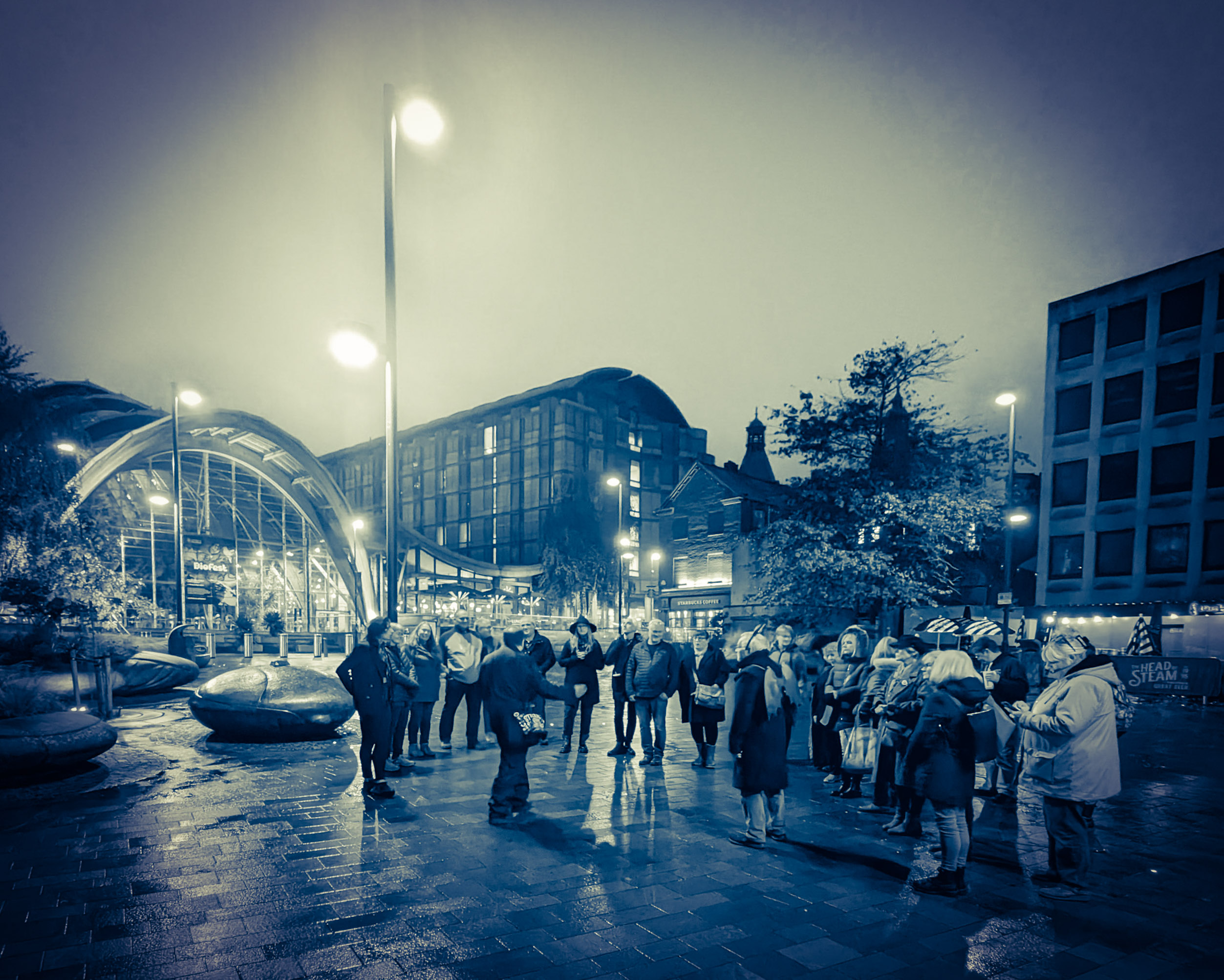 A group of people gathered round a tour guide, in Tudor Square in the centre of Sheffield, at night.