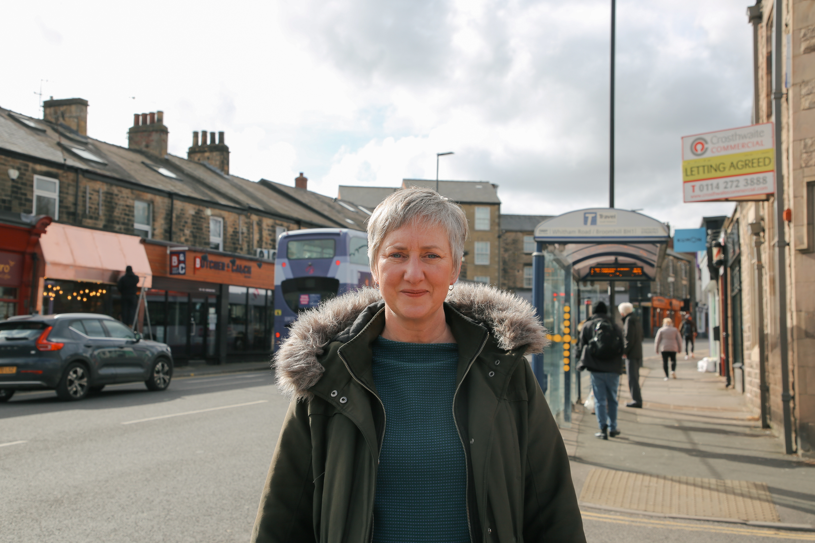 Person in a dark green jacket with a fur-lined hood standing on a city sidewalk. Behind them, a purple double-decker bus approaches a bus stop where people are waiting. Urban buildings, parked cars, and pedestrians line the street under a partly cloudy sky.