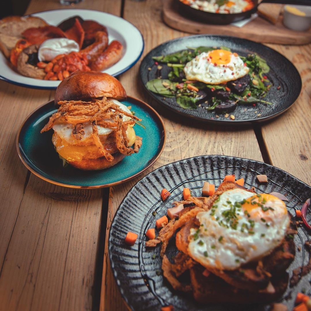 Four plates of breakfast dishes with eggs, sandwiches, greens, and a full English on a wooden table.