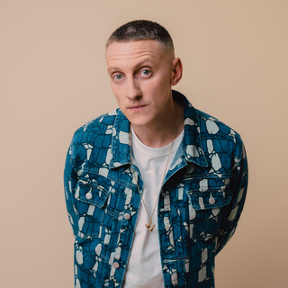 Person wearing a white T-shirt layered with a blue patterned denim jacket, standing against a plain beige background with hands behind their back.