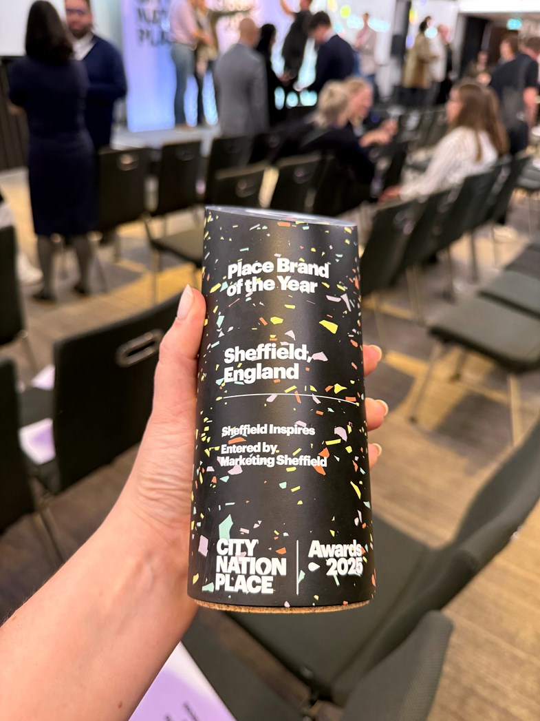 A hand holding a black totem award with white text that reads Place Brand of the Year with Sheffield, England underneath. In the background members of the conference socialise in front of a presentation stage