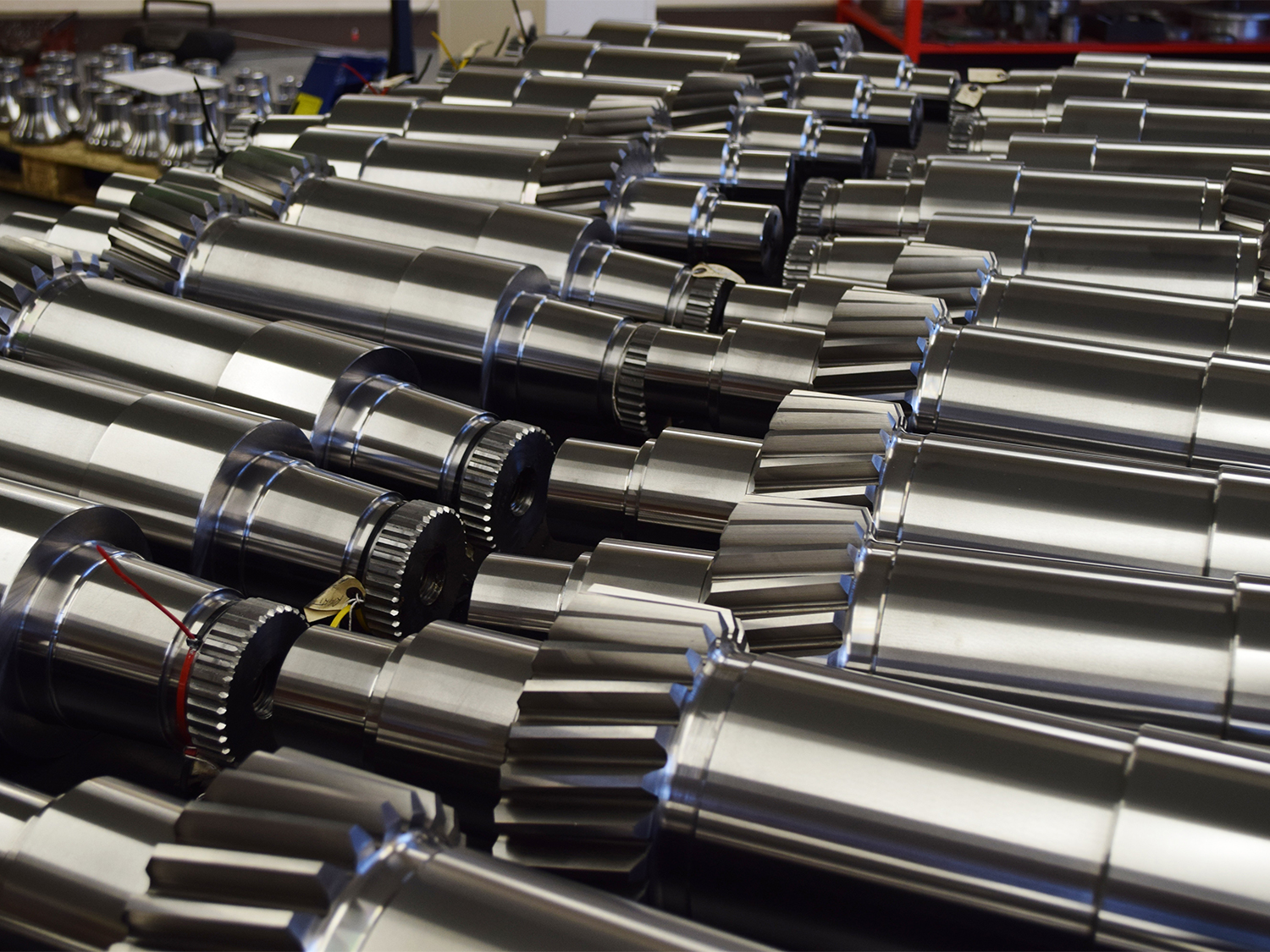 Close-up view of multiple shiny metal gears and cylindrical components arranged in rows. The parts have polished surfaces with visible teeth and grooves, suggesting precision engineering for industrial machinery or mechanical systems.