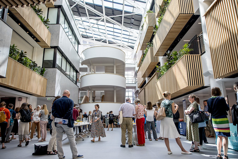 The Heartspace in Hallam University: Bright, modern atrium with a glass roof and natural light. People are gathered for an event, standing near tables with refreshments. The space features multiple levels with wooden slatted balconies, green plants, and white curved staircases in the centre, creating an open and airy atmosphere.