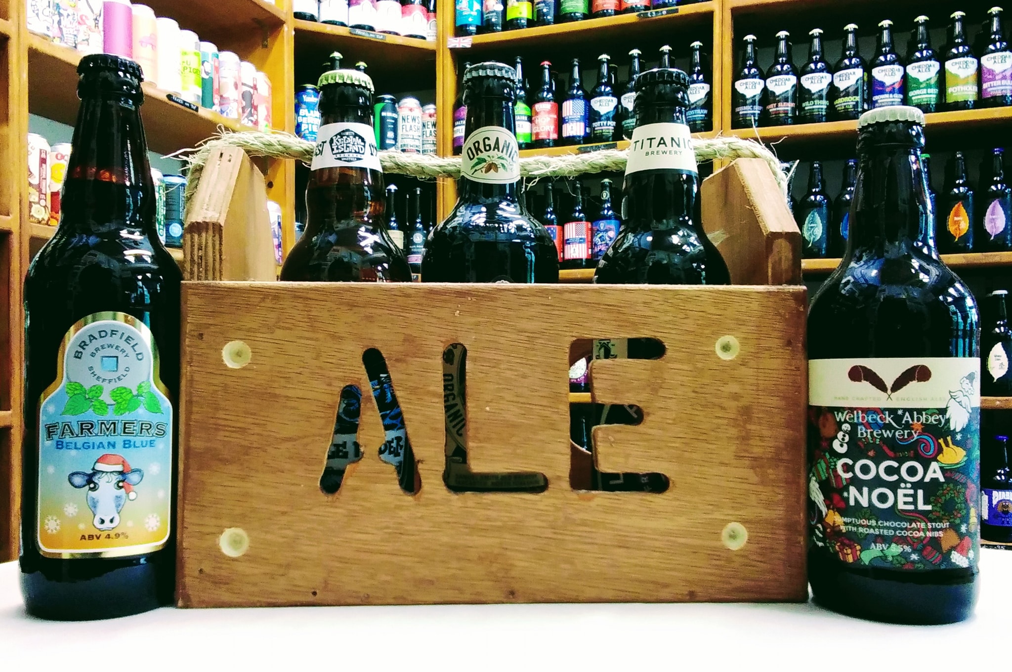 A wooden crate with the word 'Ale' cut into the side, stands on a table. It is filled with bottles of beer, and two more bottles stand on the table.