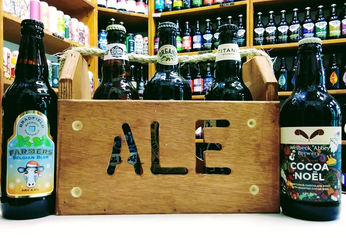 A wooden crate with the word 'Ale' cut into the side, stands on a table. It is filled with bottles of beer, and two more bottles stand on the table.