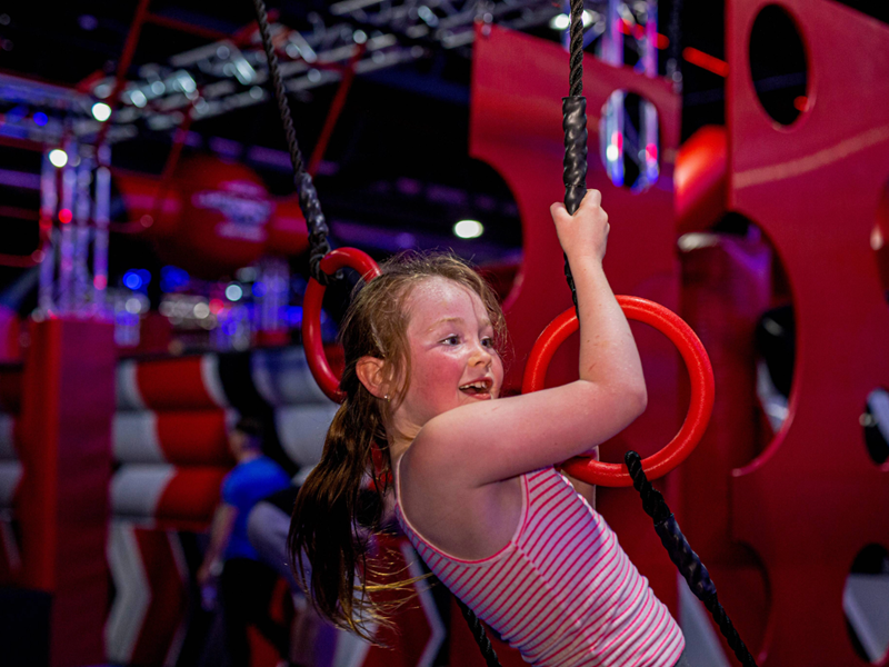 Child wearing a pink and white striped sleeveless top swings on black ropes with red rings in an indoor obstacle course featuring red and gray padded structures and bright overhead lighting.