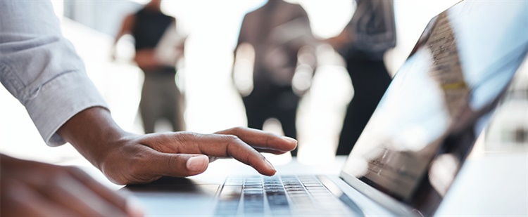 A stock image of hands typing on a laptop keuboard with three blurry figures in the background.