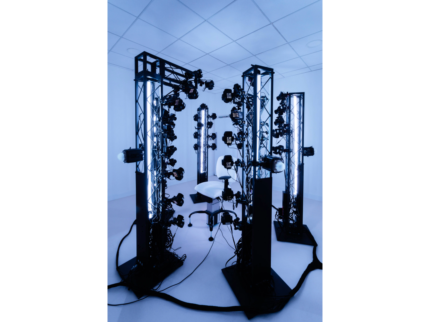 High-tech scanning setup in a white room featuring multiple vertical metal frames equipped with cameras and LED lights, arranged around a central white chair.