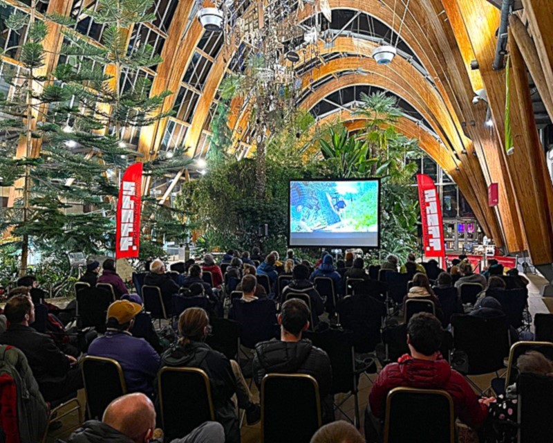 Audience seated in rows watching a film projected on a large screen inside Sheffield Winter Garden, surrounded by tall green plants and wooden arched beams. Two red vertical banners with white text flank the screen.